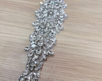 May include: A long, narrow applique featuring a dense arrangement of clear, faceted rhinestones in various shapes, including round, marquise, and pear cuts, set in a silver-toned metal base, creating a highly reflective and intricate decorative pattern.