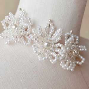 Könnte beinhalten: Zwei weiße Perlenblumenbroschen mit einer großen Perle in der Mitte jeder Blume. Die Broschen sind an einem weißen Stoffhintergrund befestigt.