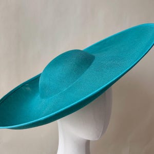 K&ouml;nnte beinhalten: Ein t&uuml;rkisfarbener Sonnenhut mit breiter Krempe und rundem Oberteil. Der Hut besteht aus einem gewebten Material und wird auf einem wei&szlig;en Schaufensterkopf pr&auml;sentiert. Die Farbe des Hutes ist ein leuchtendes, einfarbiges T&uuml;rkis.