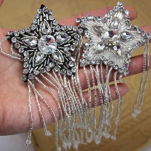 Op de afbeelding: Twee zilveren stervormige kralenpatches met franjes. De sterren zijn versierd met strass. Een ster is zwart en de andere is wit.