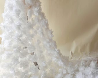 May include: White fabric with three-dimensional floral embellishments. The fabric appears to be tulle or a similar sheer material, with numerous small, white flowers densely covering the surface. The flowers are clustered, creating a textured, layered effect.