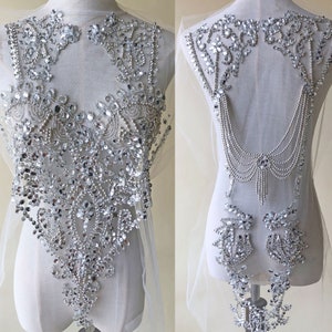 May include: A silver beaded and rhinestone embellishment for a wedding dress or other formal attire. The embellishment is shown on a white dress form and features a large, ornate design with a cascading chain of beads.