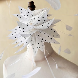 Può includere: Un affascinante copricapo di piume bianche con pois neri, disposto in un design floreale a strati. Delicate piume bianche si estendono verso l'esterno, creando un copricapo stravagante ed elegante. Il copricapo è esposto su un manichino.
