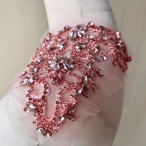 May include: A delicate pink tulle fabric with a rose gold beaded embellishment. The embellishment features a floral design with rhinestones and small beads.