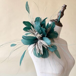 Puede incluir: Un fascinador de plumas verde azulado y blanco, exhibido en una cabeza de maniqu&iacute;. El tocado presenta grandes plumas en capas verde azulado con acentos blancos y marrones, y delicados mechones que se extienden hacia afuera. Este accesorio es adecuado para ocasiones especiales.