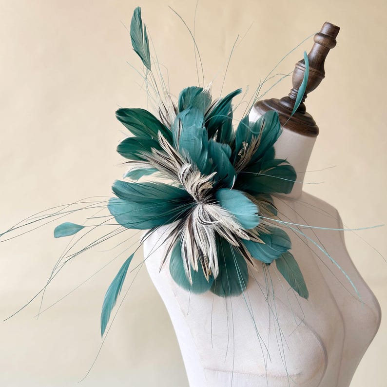 Puede incluir: Un fascinador de plumas verde azulado y blanco, exhibido en una cabeza de maniqu&iacute;. El tocado presenta grandes plumas en capas verde azulado con acentos blancos y marrones, y delicados mechones que se extienden hacia afuera. Este accesorio es adecuado para ocasiones especiales.