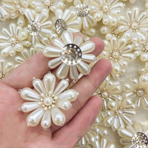 May include: A close-up of a hand holding a collection of pearl flower embellishments. The embellishments are made of white pearls and have a metal center with a decorative design. The embellishments are perfect for adding a touch of elegance to any project.