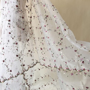 May include: White sheer fabric adorned with intricate embroidery of small burgundy flowers and green stems. The floral pattern is evenly spread across the fabric, creating a soft, romantic look. The edges feature a detailed floral border.