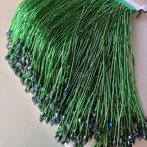 May include: A close-up of a green beaded fringe trim with iridescent green beads. The fringe is made of small, round beads that are strung together on a thin wire.