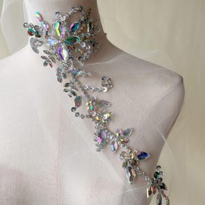 May include: A delicate, iridescent crystal applique featuring a floral design. The applique is made of small, faceted crystals that are sewn onto a mesh backing. The crystals are arranged in a pattern of leaves and flowers, creating a beautiful and elegant design.