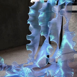 Puede incluir: Tela azul translúcida con un borde con volantes, creando un patrón ondulado. El material tiene un brillo iridiscente sutil, que atrapa la luz y crea un efecto brillante. La tela está suspendida, lo que sugiere un elemento decorativo o una instalación.