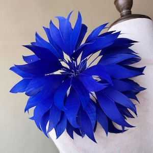 May include: A vibrant blue feather flower, possibly a brooch or decorative accent, is displayed on a mannequin. The feathers are arranged in a radial pattern, creating a full, textured appearance. The deep blue color is striking against the neutral background.