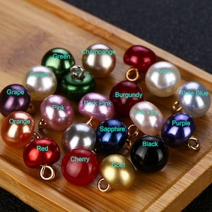 Op de afbeelding: Assortiment van 16 parelmoerachtige knopen in verschillende kleuren, waaronder groen, rood, blauw, paars, roze, oranje, goud, zwart, crème, ivoor en champagne. Elke knoop heeft een kleine gouden lus.