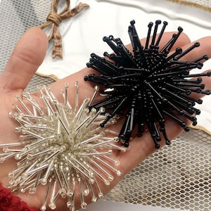May include: Two decorative beaded starburst shapes, one black and one silver, with a 3D design. The starbursts are made of small beads strung together to create long, thin spikes.