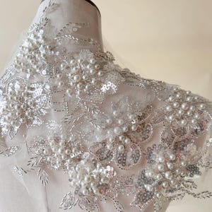 May include: A delicate white lace fabric with pearl and sequin embellishments. The fabric features a floral design with clusters of pearls.