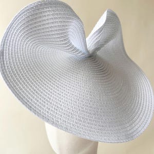 May include: A wide-brimmed white hat with a textured, woven straw-like construction. The brim features a distinct, asymmetrical upward curve on one side, creating a sculptural, undulating silhouette with concentric circular patterns throughout.
