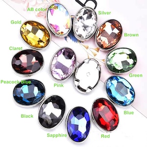 Op de afbeelding: Assortiment van ovale, gefacetteerde glazen strasssteentjes in verschillende kleuren, waaronder goud, zilver, bruin, groen, blauw, rood, zwart, roze, paars en helder. De strasssteentjes zijn gezet in zilverkleurige metalen settings.