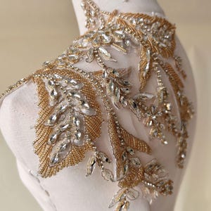 Pode incluir: Aplique de contas douradas e cristal transparente com um design floral. O aplique está sobre um fundo de tecido transparente.