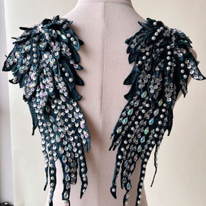 Peut inclure: Deux ornements d'épaule en forme d'ailes, de couleur vert foncé. Chaque aile est ornée de strass irisés et de petites perles blanches. Les ailes sont faites d'une matière semblable à du velours et sont exposées sur un mannequin.