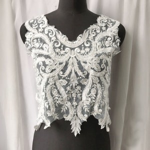 May include: A white lace top with an intricate floral and scrollwork design. The sleeveless top features a V-neckline and is displayed on a black mannequin. The delicate lace is detailed with leaves and flowers.