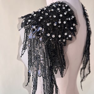 Peut inclure: Embellissement d'épaule noir en perles et paillettes avec des détails de franges. L'embellissement est sur un mannequin blanc.