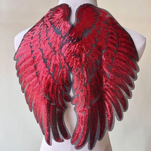 Puede incluir: Alas de ángel de lentejuelas rojas con respaldo de malla negra. Las alas son grandes y detalladas, con un diseño de plumas.