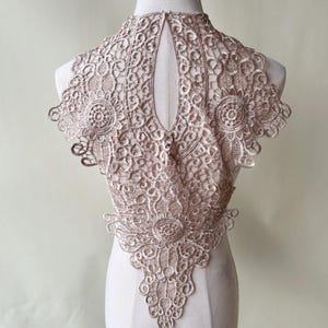 Op de afbeelding: Een delicate, beige kanten kraag met een gegolfde rand en een sleutelgat opening aan de achterkant. De kant is ingewikkeld en heeft een bloemenmotief.