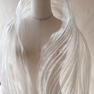 May include: A white, semi-sheer, lightweight wrap or shawl featuring prominent vertical pleats and folds. The fabric has a subtle, shimmering texture and is draped over a mannequin, creating a voluminous, layered appearance with flowing lines.