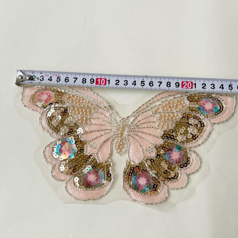 May include: A decorative butterfly applique, with a light pink and gold sequined design and iridescent accents. The applique measures approximately 15 cm wide, suitable for embellishing clothing or accessories.