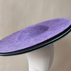 Op de afbeelding: Een lavendelkleurige hoed met een brede rand en een afgeronde kroon. De rand heeft gelaagde zwarte, witte en blauwgroene accenten. De hoed wordt getoond op een witte mannequinhoofd, wat de vorm en het ontwerp benadrukt.