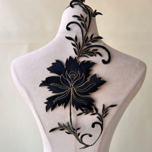 Può includere: Applique floreale nera con ricamo dorato. L'applique presenta un grande fiore con un design dettagliato e foglie intricate.