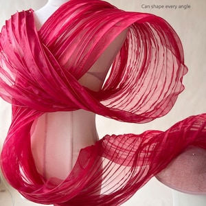 Puede incluir: Un envoltorio de tela plisada, transparente y de color rojo vibrante, drapeado sobre un maniquí. El material presenta un diseño estructurado y en capas con líneas horizontales visibles. El texto 'Can shape every angle' aparece en la esquina superior derecha.