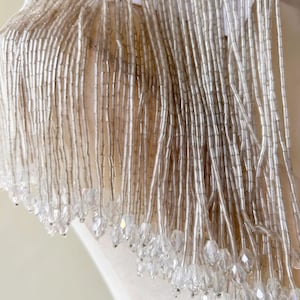 May include: Close-up of a white garment with intricate beaded fringe. The fringe is composed of long, thin, silver beads and larger, clear crystal-like beads at the bottom, creating a shimmering effect. The garment appears to be a shoulder or neckline detail.
