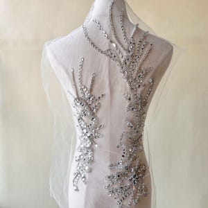 Op de afbeelding: Zilveren pailletten- en strass-applicatie op transparante stof. Het ontwerp heeft een symmetrisch, bloem geïnspireerd patroon met sprankelende versieringen. Ideaal voor het verfraaien van kleding of accessoires.