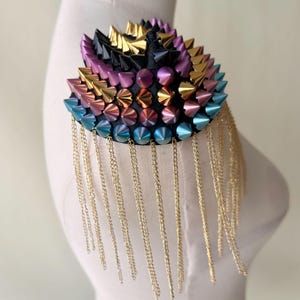 Peut inclure: Une épaulette colorée à plusieurs niveaux avec des franges de chaîne dorées. La pièce est faite d'une base en tissu noir avec des rangées de pointes métalliques dans des tons d'or, de violet, de rose, de bleu et de turquoise.