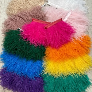 May include: A collection of colorful feather trim in various shades, including pink, green, orange, yellow, blue, and purple. The feathers are arranged in layers, showcasing their texture and vibrant hues. Some trims have a red ribbon.