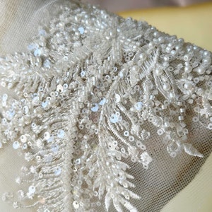 May include: Close-up of a white beaded and sequined fabric trim. The trim features a delicate floral design with small pearls and sequins.