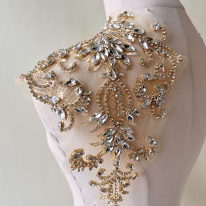Puede incluir: Una delicada aplicación de oro y cristal con un diseño floral. La aplicación está hecha de una tela transparente con un bordado de hilo dorado y diamantes de imitación transparentes.