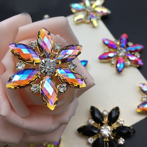 Pode incluir: Conjunto de enfeites de strass em forma de flor iridescentes com engastes dourados. Os strass têm um efeito arco-íris e são rodeados por strass transparentes mais pequenos.