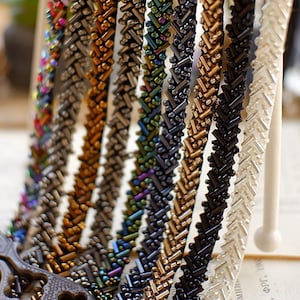 Puede incluir: Una colección de ocho tiras de adorno de cuentas en varios colores, incluyendo plata, oro, negro, iridiscente y transparente. El adorno está hecho de cuentas pequeñas, espaciadas uniformemente, dispuestas en un patrón de chevron.