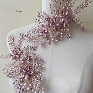 Op de afbeelding: Een paar decoratieve bloemenversieringen. Elk stuk heeft een cluster van roze kristallen bloemen en heldere kristallen accenten, gezet op een goudkleurig draadframe. Het ontwerp bevat bladachtige details, wat een elegante, sierlijke uitstraling creëert.