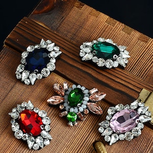 May include: Five different coloured rhinestone embellishments with a variety of shapes and sizes. The embellishments are on a wooden surface.