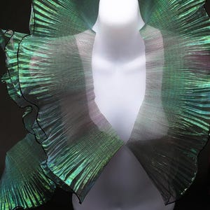 Puede incluir: Un collar verde iridiscente con bordes con volantes y ribete negro. El collar está drapeado sobre un maniquí blanco, mostrando su textura plisada y su diseño dramático. El color del collar cambia de verde a negro.