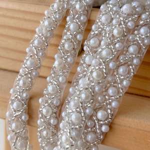 May include: A decorative trim featuring white faux pearls and silver seed beads arranged in a repeating geometric pattern on a sheer white mesh ribbon. The intricate beadwork creates a textured, raised surface across the length of the fabric trim.