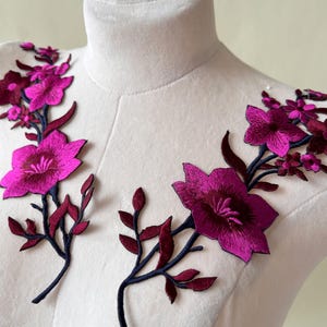May include: Embroidered floral appliques in shades of fuchsia and burgundy. The flowers have detailed stitching and are attached to dark blue stems with burgundy leaves. The appliques are displayed on a white mannequin.