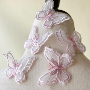 May include: Six pink butterfly appliques made from a sheer, delicate fabric. The butterflies feature a pink embroidered design and are ideal for adding a touch of whimsy to clothing or crafts.