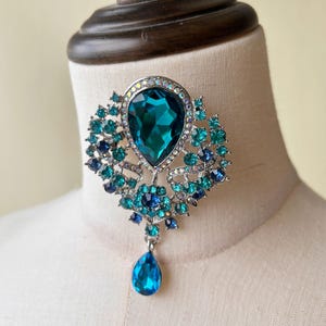 May include: A silver and blue jeweled pendant with a large teardrop-shaped blue stone. The pendant is on a silver chain and has a smaller teardrop-shaped blue stone hanging from the bottom.