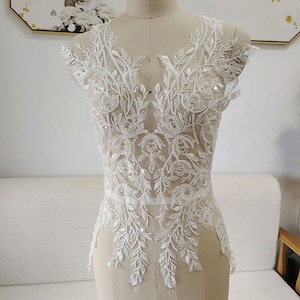 May include: White lace applique featuring beaded floral and leaf designs. The applique is displayed on a white mannequin and is ideal for adding a touch of elegance to a wedding dress or other special occasion garment.