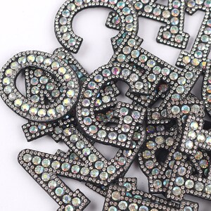 Puede incluir: Un mont&oacute;n de letras de fieltro negro cubiertas de diamantes de imitaci&oacute;n iridiscentes. Las letras son de diferentes tama&ntilde;os y formas, y est&aacute;n dispuestas en un orden aleatorio.