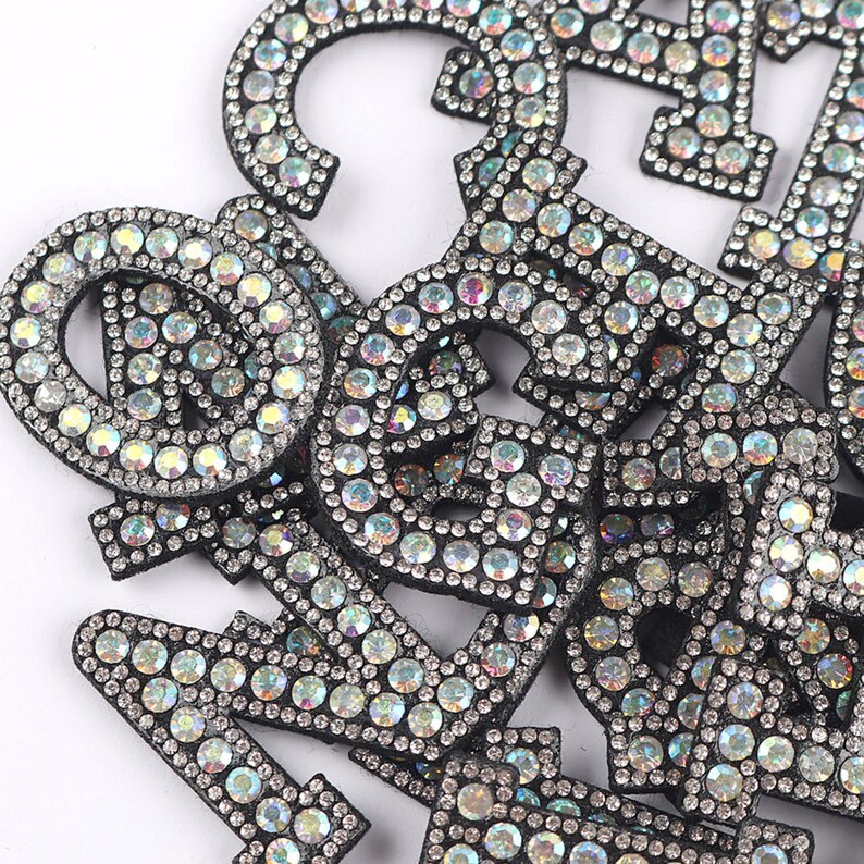 Puede incluir: Un mont&oacute;n de letras de fieltro negro cubiertas de diamantes de imitaci&oacute;n iridiscentes. Las letras son de diferentes tama&ntilde;os y formas, y est&aacute;n dispuestas en un orden aleatorio.